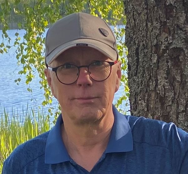 Hannu Mars kesävaatteissa ja lippalakissa. Taustalla kaunis suomalainen järvimaisema ja koivu