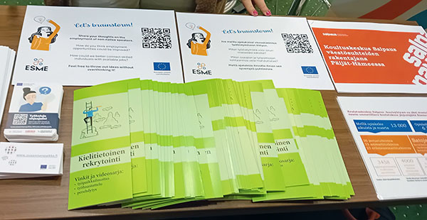 Erilaisia mainoslehtisiä pöydällä, teksti kielitietoinen rekrytointi.