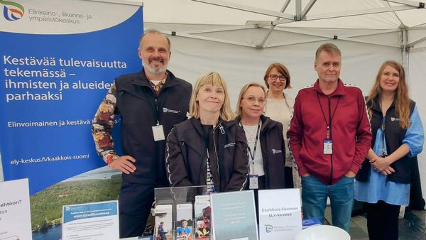 Kuusi henkilöä seisoo vierekkäin tapahtumateltassa, esitteitä ja juliste.