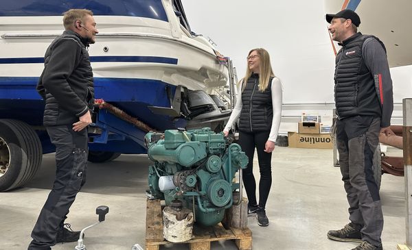 Kolme henkilöä hallissa, jossa näkyy veneitä ja veneen moottori.