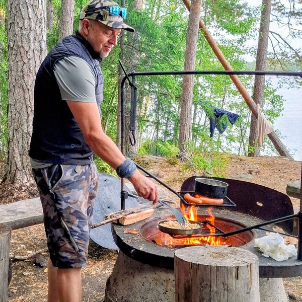 Iäkäs mies paistaa makkaraa retkipaikan nuotiossa järven rannalla.
