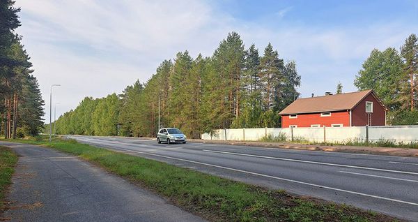 Henkilöauto ajaa valtatiellä 20, taustalla punainen talo ja sen edessä korkea aita.