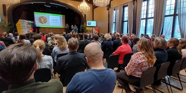 Henkilöt istuvat rivissä auditorion lavalla.