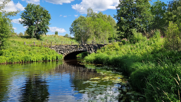 Vanha kivinen silta joen yli vehreässä maalaismaisemassa.