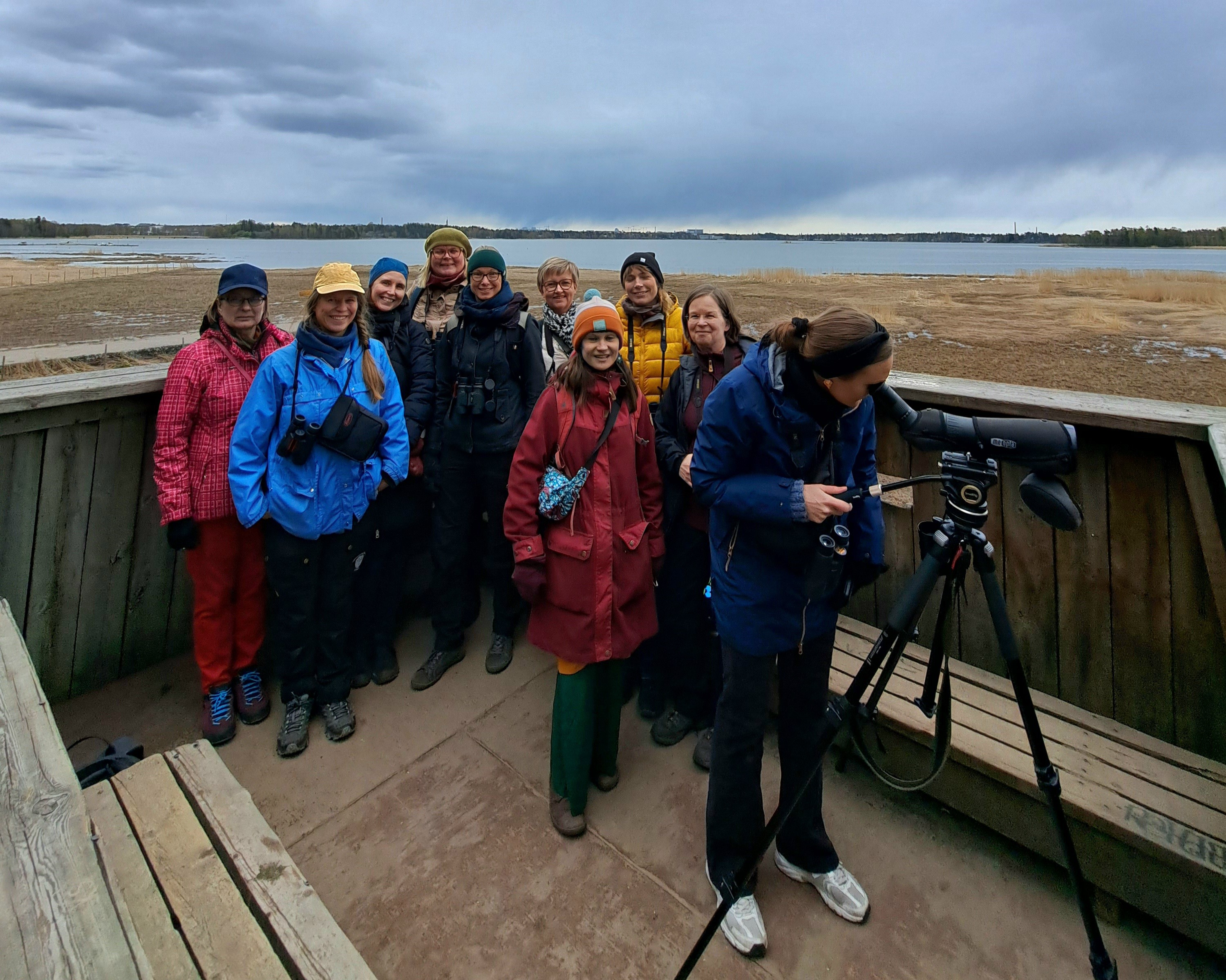 Ryhmä ihmisiä seisoo lintutornin laella katsoen kameraan. Etualalla yksi henkilö tarkkailee kaukoputkella lintuja.