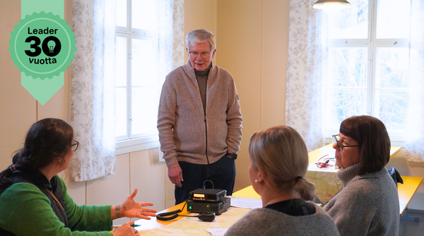 Ihmisiä Alastarolla pöydän ääressä. Keskellä pöytää kyläradio.