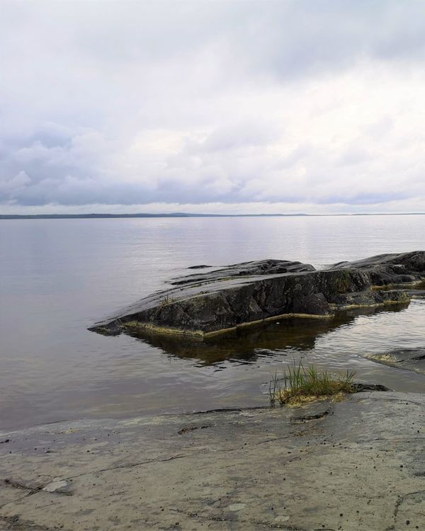 Rannalta kuvattu merimaisema. Meri on tyyni ja taivas pilvinen. Rannan edustall on kalliota.
