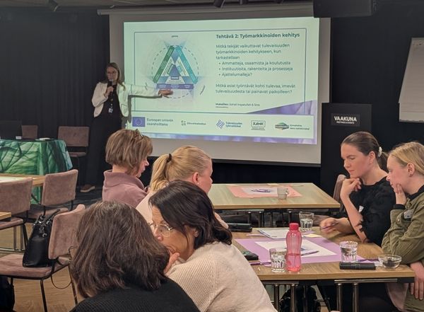 KaakkoACT projektipäällikkö Kati Viljakainen antamassa työpajaohjeistusta lavalla. Kuvan etualalla pohdiskelevia asiantuntijoita tehtävien äärellä.