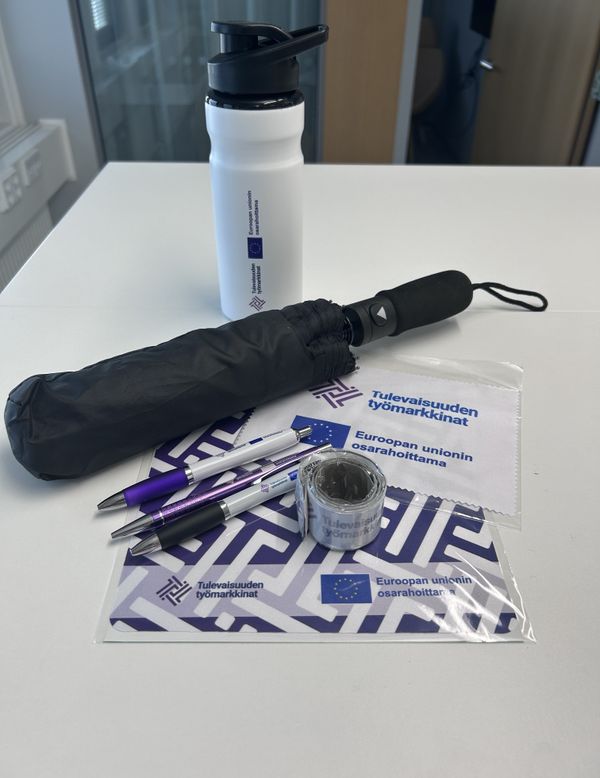 kuva tt-hankkeen tuotepaketista, juomapullo, sateenvarjo, mikrokuituliina, heijastin, hiirimatto ja kolme kynää