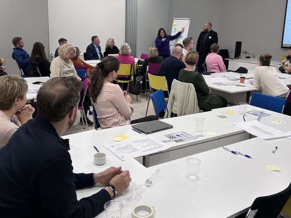 kuva kokoustilasta, jossa ihmiset istuvat valkoisten pöytien ääressä ja kuuntelevat edessä olevaa puhujaa